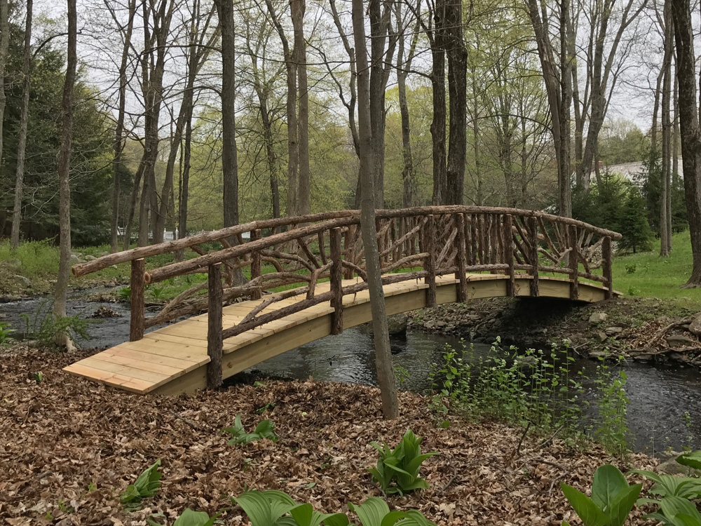 Rustic bridge crossing a stream built using bark-on trees and branches titled Storm's Crossing