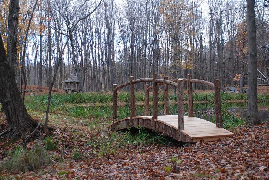 Rustic bridge near a pond custom built using cedar trees and branches titled the Red Hook Watershed Bridge